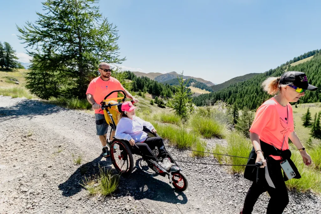 Une jeune fille en fauteuil tout-terrain Hippocampe en randonnée à Valberg, accompagnée de ses parents.