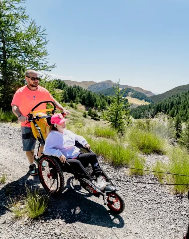 Une jeune fille en fauteuil tout-terrain Hippocampe en randonnée à Valberg, accompagnée de ses parents.